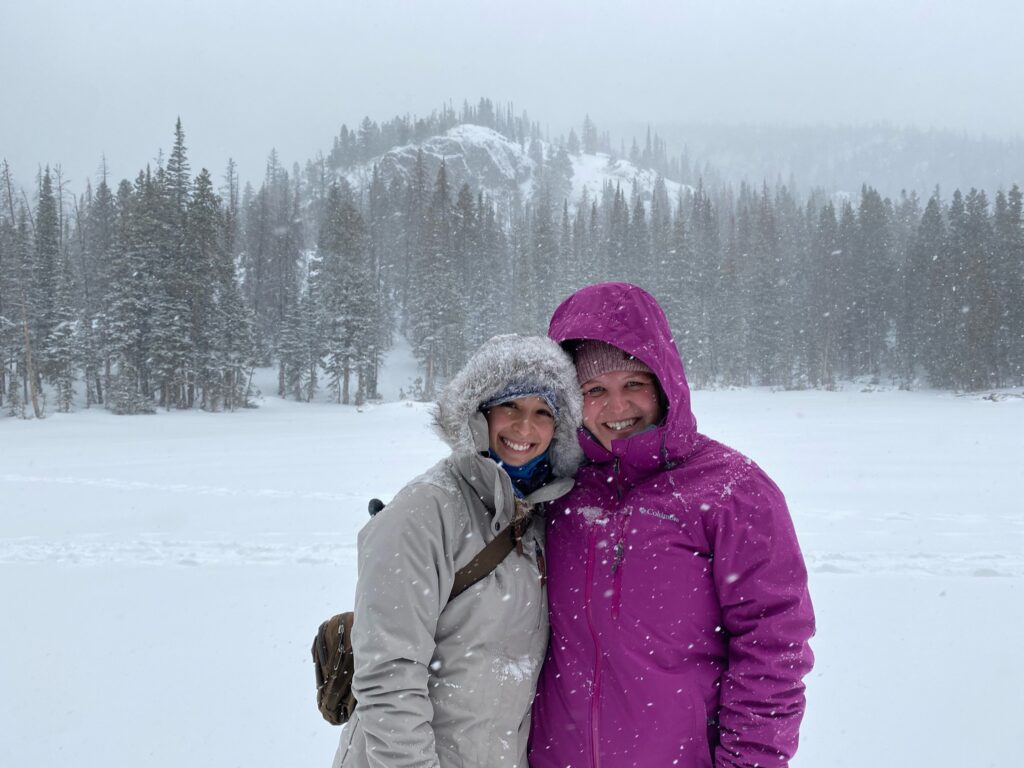 Two women in winter jackets standing in a snowy field with snow capped mountains and trees in the background