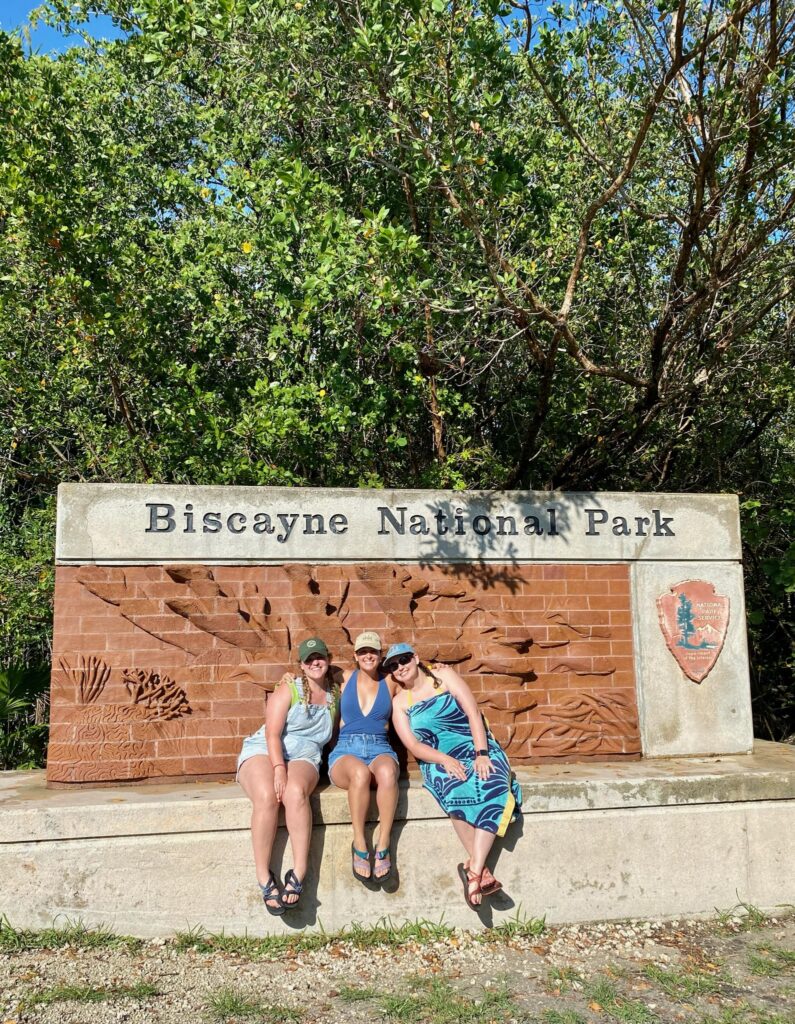 Madi (author), Lindsay (author), and their friend Megan sitting in front of the Biscayne National Park Sign. 