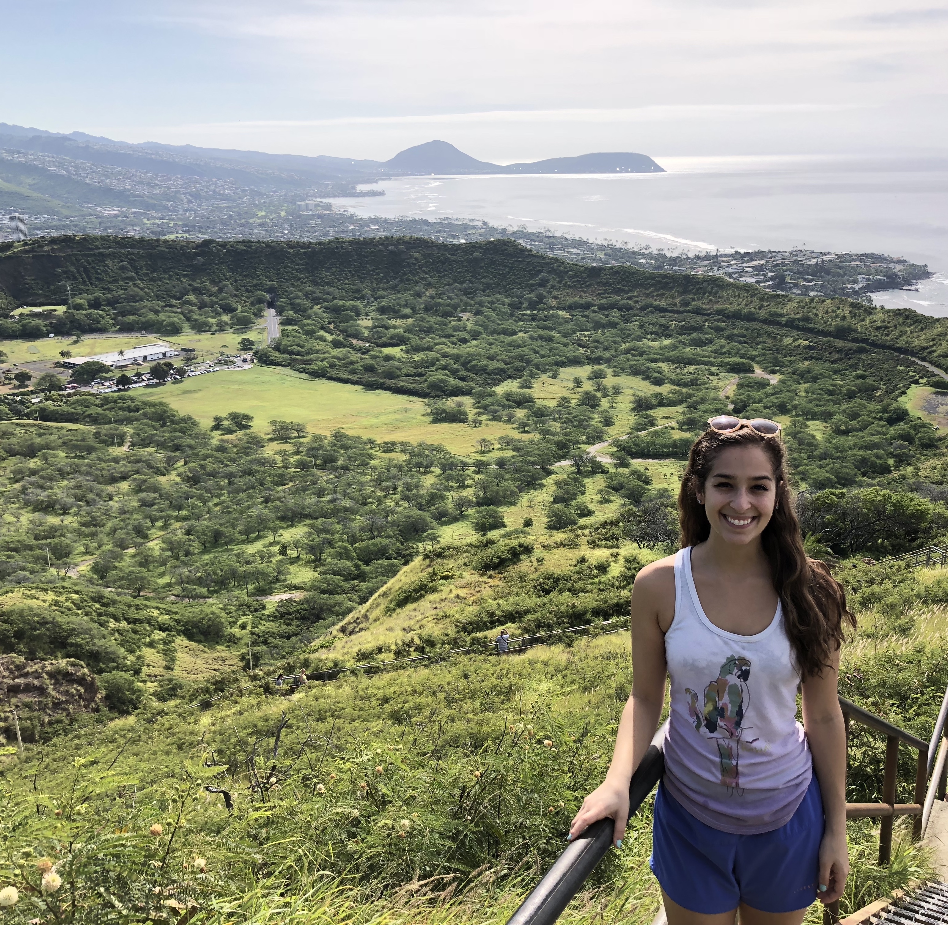 This photo shows the Diamond Head Crater, a very green crater in the Oahu landscape. You can also see the coastline and ocean in the background. There is a smiling girl posing in front of the crater.