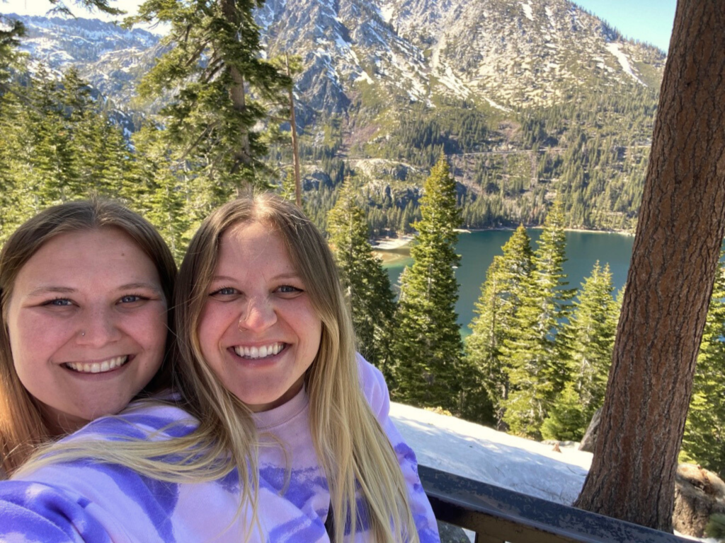 Two women (the author and her sister) standing in front of Emerald Bay, Lake Tahoe in April. The Emerald Bay has blue green water, is surrounded by evergreen trees, and is located at the base of snow capped mountains. 