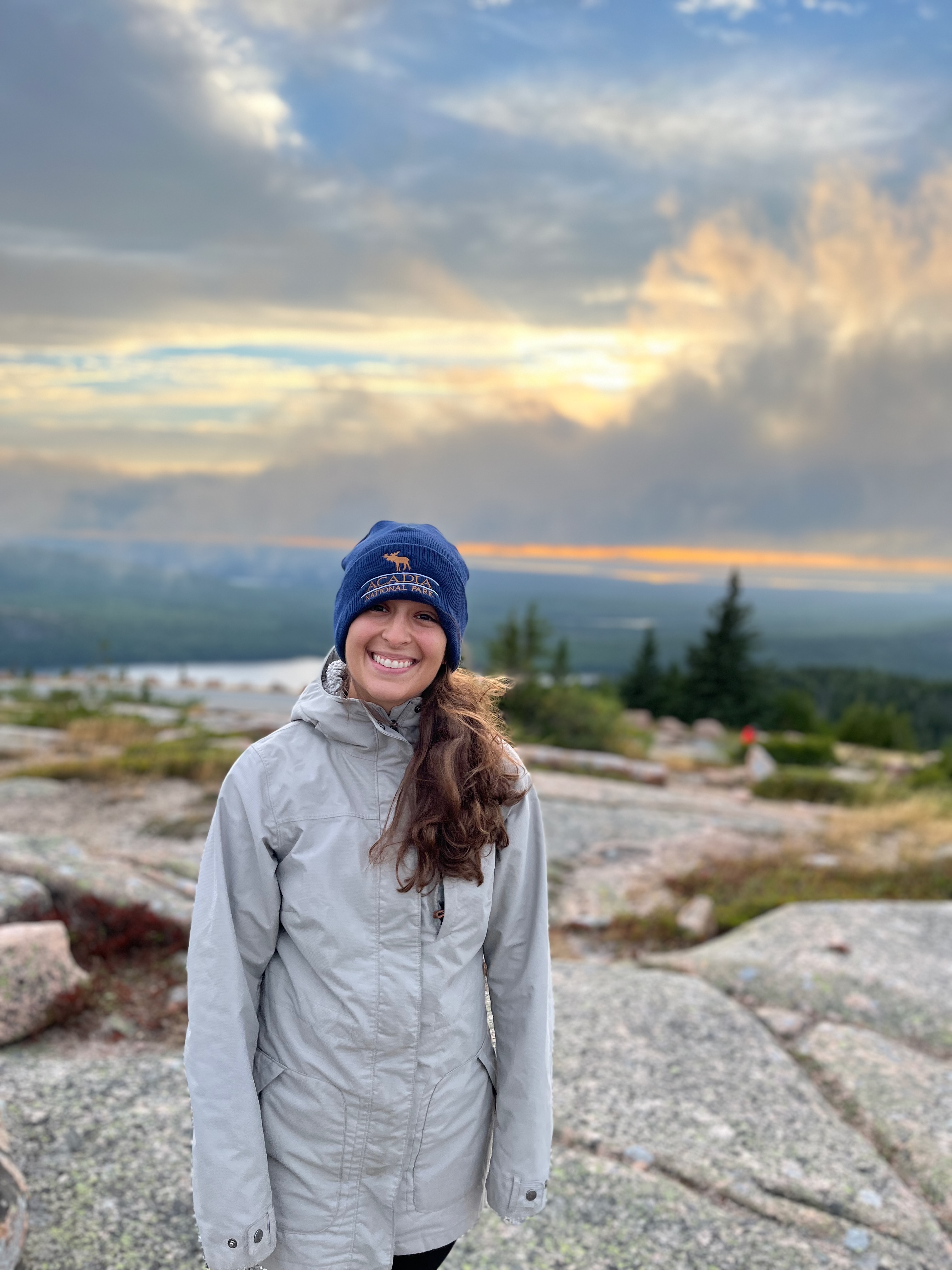 A cloudy sunset view from Cadillac mountain summit with a girl in the foreground smiling wearing an Acadia winter hat.