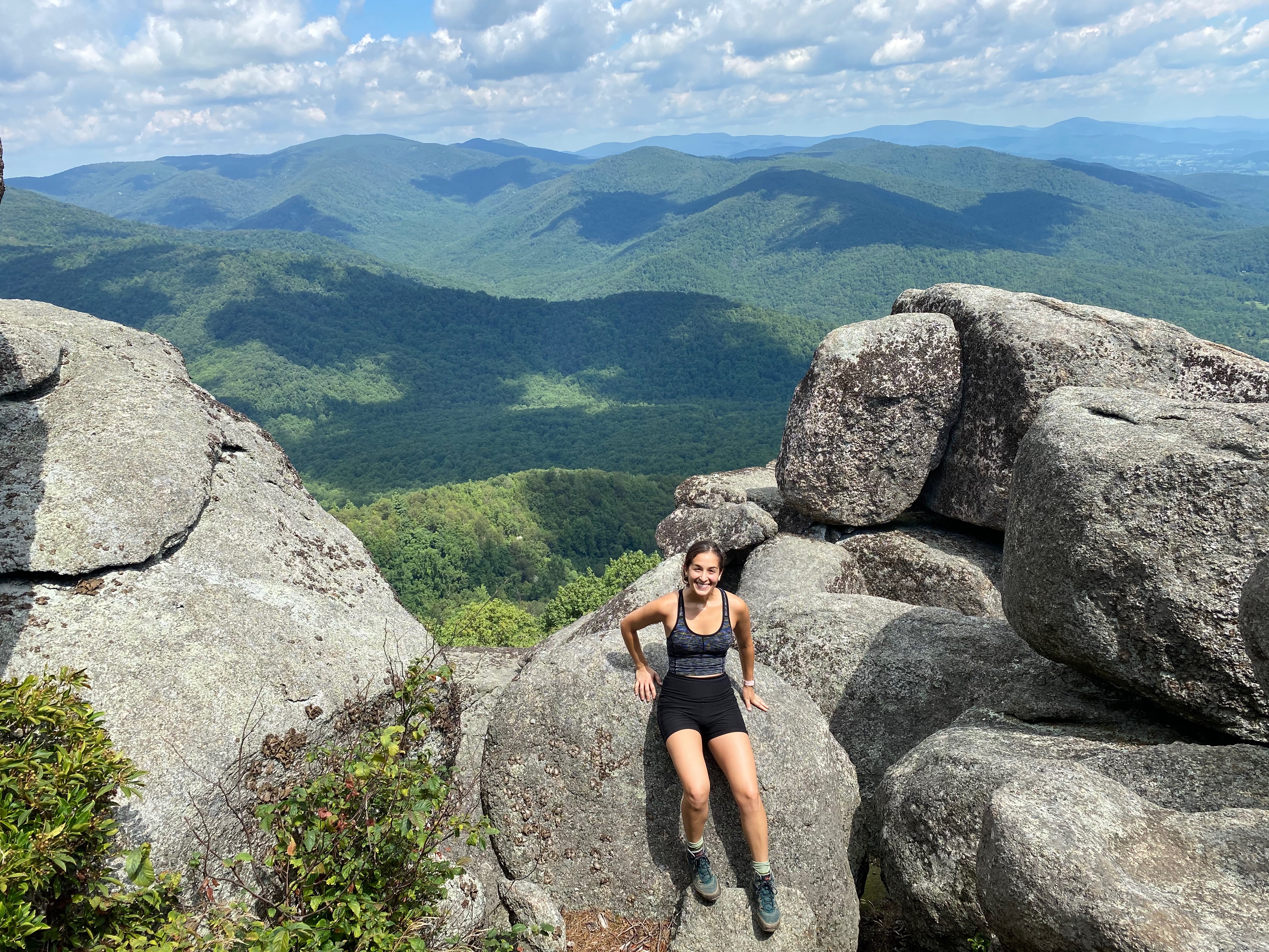 Breathtaking panoramic view from the summit of Old Rag Mountain, showcasing the rugged landscape, rocky outcrops, and lush greenery of the Shenandoah National Park. A woman sits on the rocky mountain top in the middle of the frame.