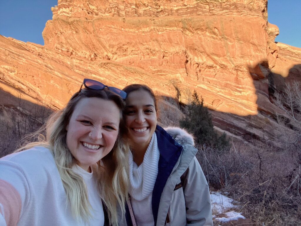 Madi and Lindsay (blog authors) standing in front of a large red rock on the Red Rocks Amphitheater hiking trail. 