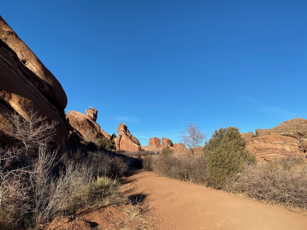 Red Rocks Amphitheater hike. Red rocks in the back with green shrubs on each side of the dirt trail. 