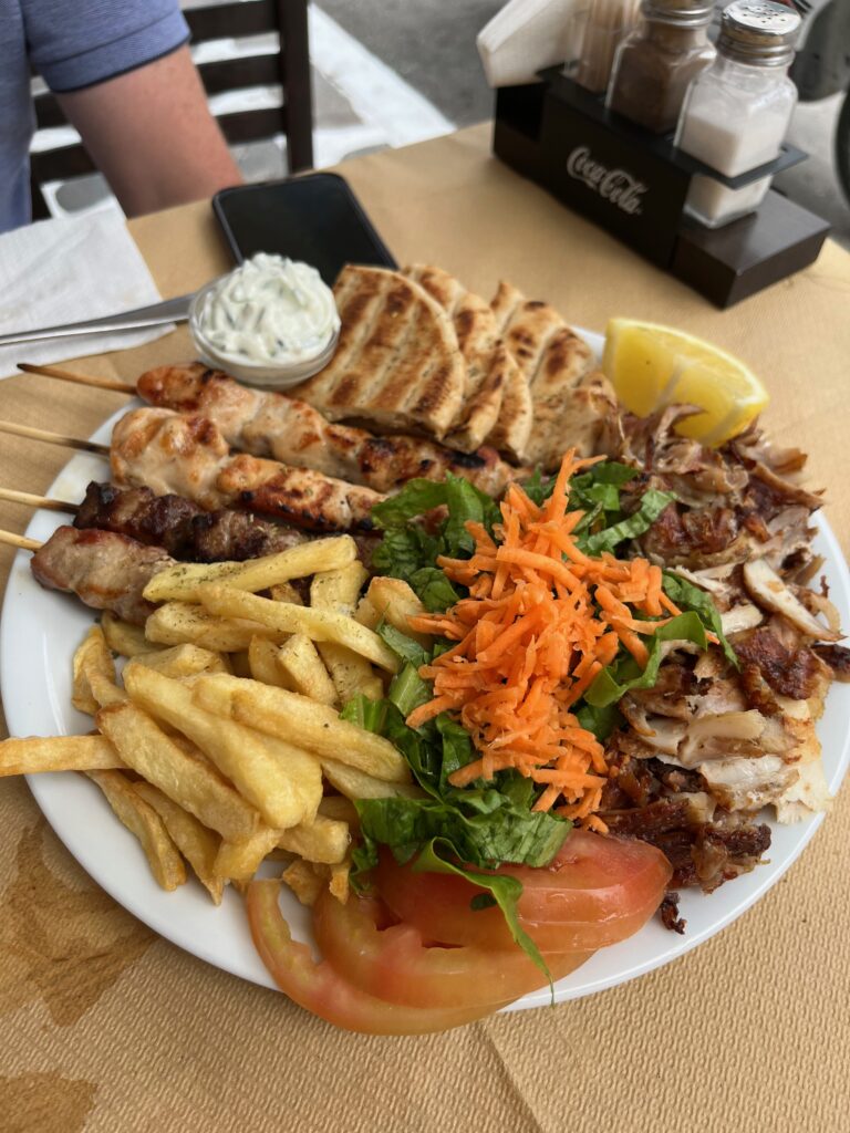 A mouthwatering display of a generous serving of Greek souvlaki on a large plate, featuring skewers of perfectly grilled meat, accompanied by vibrant vegetables, drizzled with savory sauces, and garnished with fresh herbs.