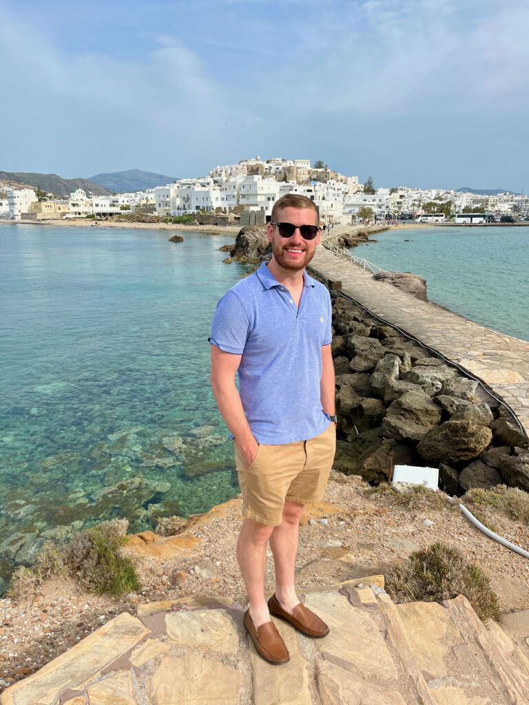 Man standing happily with a captivating backdrop of Naxos town, showcasing charming white-washed buildings and the scenic beauty of the coastal Greek town.