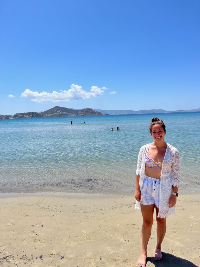 A woman standing in the foreground against the stunning backdrop of Agios Georgios Beach in Naxos. The golden sand stretches along the shoreline, meeting the turquoise waters of the Aegean Sea, while the woman takes in the beauty of the coastal paradise.