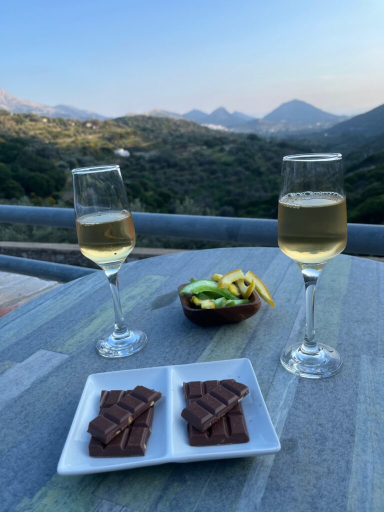 Romantic indulgence with a view: Two champagne flutes filled with effervescent bubbles, surrounded by tempting candied fruits and decadent chocolate, set against the enchanting backdrop of the Naxos countryside. A perfect blend of luxury and natural beauty, capturing the essence of a delightful moment in this idyllic Greek island setting.