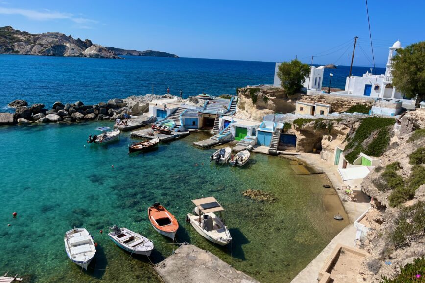 Idyllic coastal scene in Mandrakia, Milos, Greece, featuring traditional fishing boats gently bobbing in the crystal-clear turquoise waters. Mandrakia is a MUST on your Milos Itinerary.