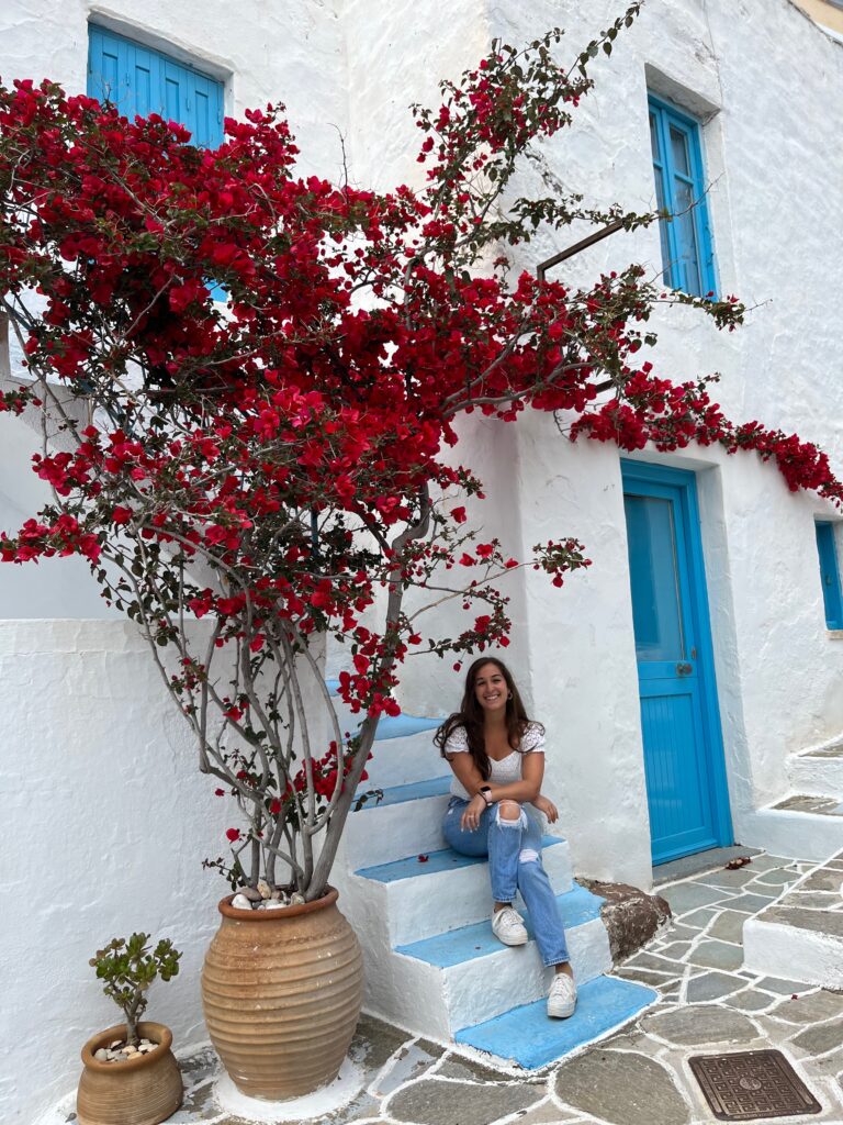 Woman seated on the steps of a stunning Greek white and blue building, framed by a vibrant red floral tree in full bloom