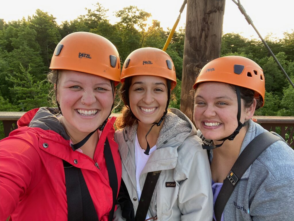 Madi, Lindsay, and their friend Megan in orange hard helmets on a zip lining platform in the Great Smoky Mountains. 